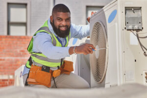 A black man kneels next to an air conditioning unit with a screen driver in place to remove the siding with a smile on his face, ready to perform the HVAC repair.